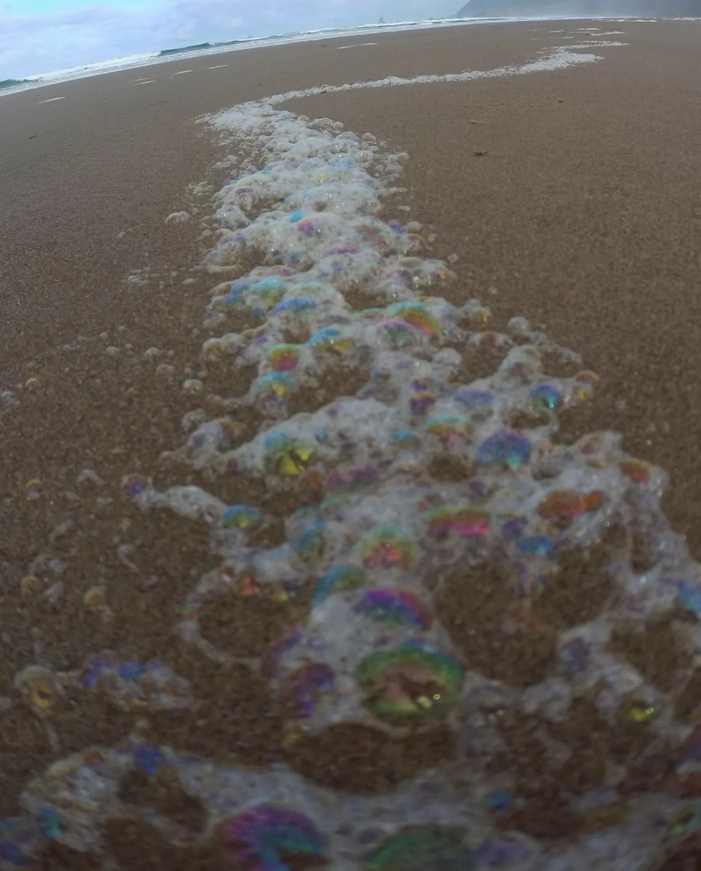 A line of sea foam and iridescent bubbles trailing from the near sand back toward the surf