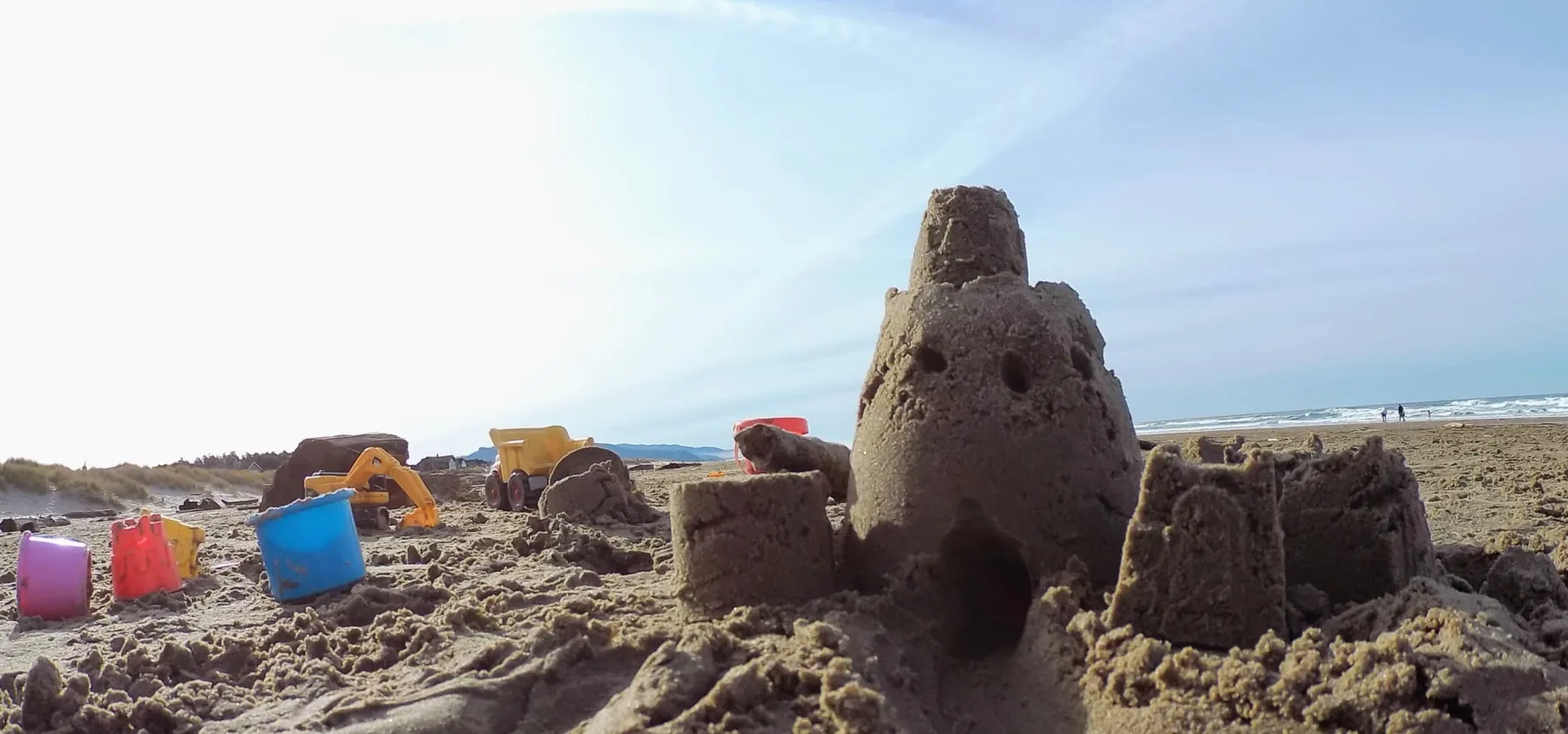 A hand-built sandcastle on a wide beach, buckets and toy trucks scattered nearby, the surf in the distance
